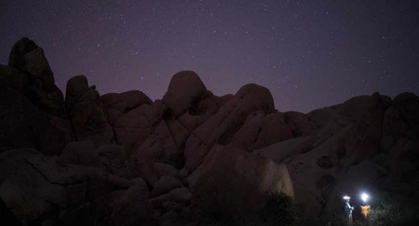 A headlamp illuminates a person sitting at the bottom of a large rock formation, resting under a purple night sky dotted with stars